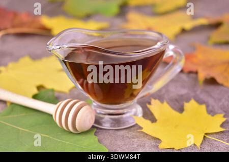 Süßer Ahornsirup in einem Glas Soße Boot und Ahornblätter auf grauem Hintergrund. Stockfoto