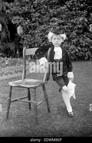 Viktorianisches Kind in georgianischer Tracht posiert neben einem Holzstuhl in einem Garten Stockfoto