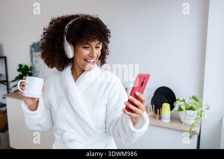 Glückliche junge Frau im Bademantel, die Online-Musik mit kabellosen Kopfhörern genießt Stockfoto