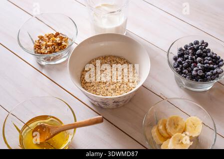 Hafer in einer Keramikschale umgeben von Milch, Nüssen, gefrorenen Heidelbeeren und Honig, gesunde Frühstückszutaten bereit für hausgemachte Porridge Zubereitung Stockfoto