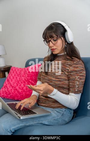 Junge Frau mit Kopfhörern und Brillen, die von der Couch aus arbeitet und auf einem Laptop schreibt Stockfoto