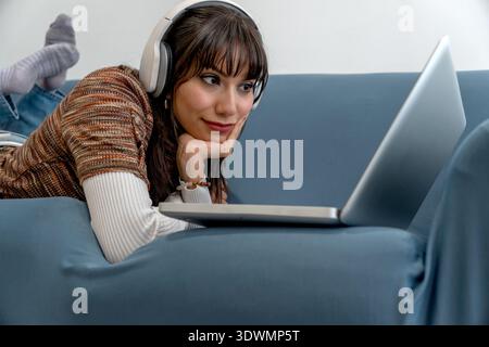 Frau, die auf dem Sofa liegt und Online-Inhalte mit Kopfhörern und Laptop genießt Stockfoto