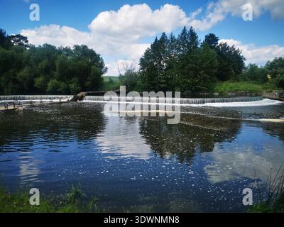 Ein kleiner Wasserfall fließt in einen reflektierenden Fluss, umgeben von üppigen Bäumen und einem hellen Himmel. Dieses Bild repräsentiert friedliche Landschaften, natürliche Schönheit, Stockfoto
