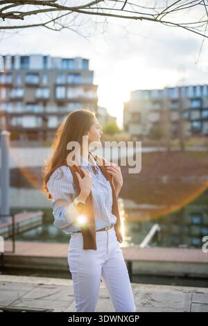 Elegante junge Frau im alten Geldstil, die während der goldenen Stunde am Flussufer in der Nähe des Prager Piers spaziert. Ruhige, luxuriöse Ästhetik und selbstbewusstes europäisches Ambiente Stockfoto
