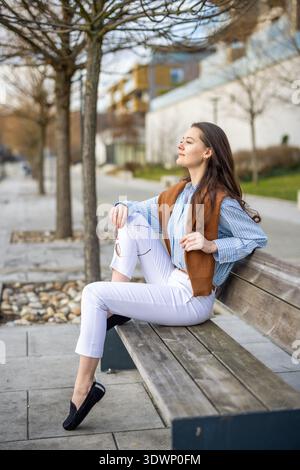 Elegante junge Frau im alten Geldstil, die auf einer Bank in der Nähe des Piers in Prag sitzt und Sonnenbrille hält und das Sonnenlicht des Frühjahrs genießt. Ruhiger Luxus Stockfoto