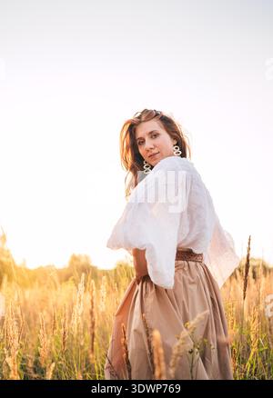 Junge Frau in weißer Bluse und beigefarbenem Rock, die frei durch goldene Wiese geht und den Sonnenuntergang genießt warmes Sonnenlicht friedliche Naturatmosphäre, die Freiheit ausdrückt Stockfoto