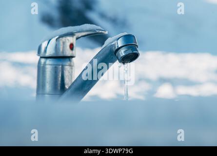 Nahaufnahme eines gefrorenen Metallhahns im Freien, der mit Schnee und Eis bedeckt ist, mit einem einzelnen Eiszapfen, der am Auslauf hängt und die Herausforderungen im Winter symbolisiert Stockfoto