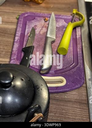 Drei Messer auf lila Schneidebrett links Midprep, rostfreies Kochmesser und Schälmesser mit gelbem Schäler und schwarzem Universalmesser, Topfdeckel R Stockfoto