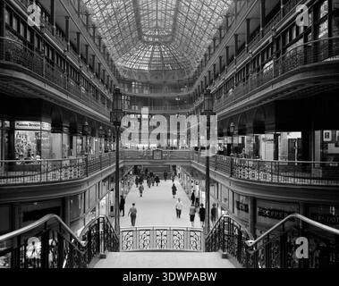 Dieses beeindruckende Schwarzweiß-Foto von Martin Linsey fängt die Pracht der Arcade in Cleveland, Ohio, ein, die 1966 in Richtung Süden in Richtung Euclid Avenue blickte. Das mehrstöckige Eisenwerk, die Glasdecke und die verschiedenen Ladenfronten des historischen Gebäudes sind zu sehen. Zahlreiche Personen durchstreifen das elegante Interieur. Stockfoto