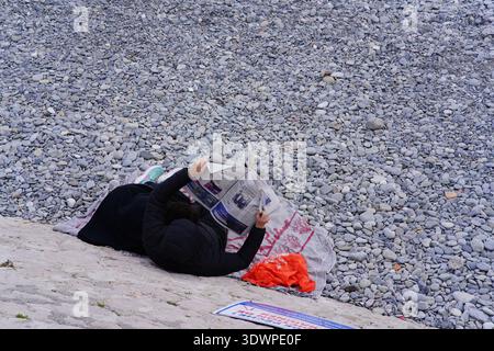 Frau, die sich am Kiesstrand in Nizza, Frankreich, entspannt und mit ihrer orangen Tasche in der Nähe eine Zeitung liest. Nizza, Frankreich Stockfoto