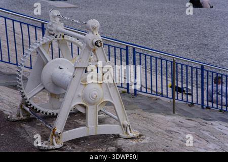 Historische weiße Seilwinde mit großen Zahnrädern an der Promenade des Anglais in Nizza, Frankreich, neben blauem Geländer und Kiesstrand. Stockfoto