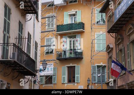 Enge Straße in Vieux Nizza mit bunten Pastellgebäuden, grünen Fensterläden, schmiedeeisernen Balkonen unter Gerüsten und einer französischen Flagge im Histori Stockfoto