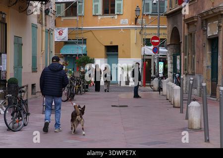 Mann, der mit seinem Hund durch eine charmante, enge Straße in Vieux Nice mit farbenfrohen Fassaden, Fahrrädern und Fußgängern in der historischen Altstadt von Nizza geht Stockfoto