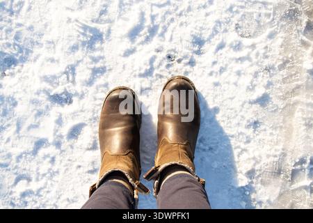 Damenfüße in braunen Winterstiefeln stehen im Schnee für die Themen Winterspaziergang, Kälteschutz, Schuhe für kaltes Wetter, rutschige Oberflächen Stockfoto