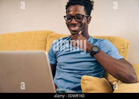 Glücklicher schwarzer Mann, der zu Hause an einem Notebook arbeitet, lächelt und gleichzeitig Online-Arbeit und digitalen Lebensstil verwaltet Stockfoto
