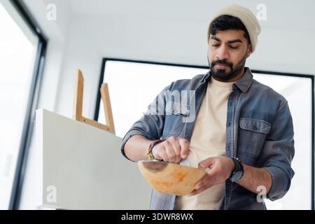 Erwachsener indischer Mann mit Creme-Mütze und Denim-Hemd, der Farbe in der Schüssel neben grundiertem Canvas umrührt. Staffelei, Studio, minimalistisch, Highkey, Tageslicht, edito Stockfoto