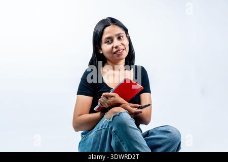 Eine fröhliche südostasiatische Frau, die auf weißem Hintergrund sitzt, ein rotes Tagebuch und einen Stift mit einem freundlichen Lächeln hält und Bildung und Freude repräsentiert. Stockfoto