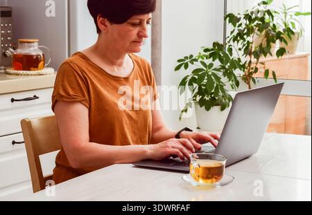 Eine reife Frau in lässiger Kleidung arbeitet an einem Holztisch mit einer Tasse Tee, umgeben von Pflanzen im Innenbereich Stockfoto