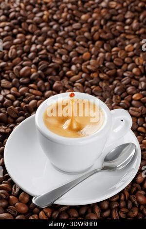 Kleine weiße Espresso-Tasse mit Spritzern und Tropfen, voller aromatischem Kaffee, der auf einem Bett aus gerösteten Bohnen liegt Stockfoto