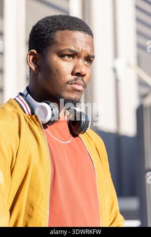Afroamerikaner, der draußen in der Nähe des Gebäudes steht, senfgelbe Jacke und Kopfhörer trägt Stockfoto