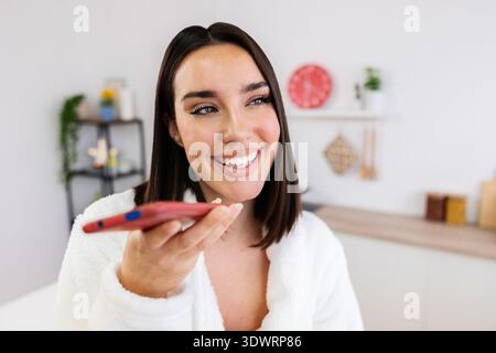 Lächelnde Frau, die den Sprachassistenten am Smartphone in der Küche benutzt Stockfoto
