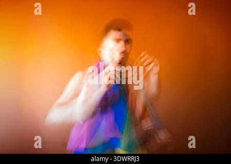 Junger Mann, der vor Bewegung unscharf steht und einen Regenbogenschal vor einem orangefarbenen Hintergrund hält Stockfoto