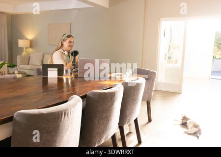Frau, die zu Hause am Esstisch sitzt und mit dem Mikrofon mit Kopfhörern spricht und Laptop benutzt. Podcaster, Fernbedienung, Creator, Innenraum, Sonnenlicht, Kat., b Stockfoto