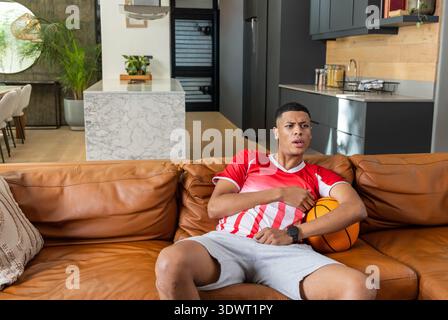 Ein Mann in seinen 20ern sitzt zu Hause auf dem Sofa und hält Basketball, trägt rotes Jersey, Kopierraum. Männlich, Sportler, lässig, modern, modern, Innenraum, offen Stockfoto