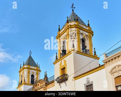 Ronda, Spanien - 2. Februar 2026: Blick auf die Glockentürme der Iglesia de Nuestra Señora del Socorro, die den klaren blauen Himmel durchstechen, geschmückt mit gelben Tri Stockfoto