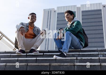 Verschiedene männliche Freunde sitzen auf Steintreppen auf der plaza und unterhalten sich mit Jacken und Rucksäcken Stockfoto