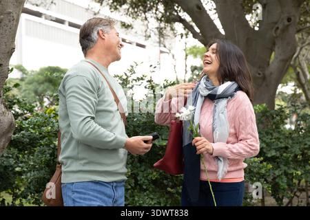 Ein Paar Senior Mann lächelt, eine Erwachsene Frau lacht in einem rosa Pullover, hält Telefon und Gänseblümchen im Park Stockfoto
