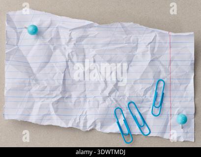 Ein zerknittertes, gerissenes Blatt liniertes Papier wird mit zwei blauen Druckstiften auf einem grauen Hintergrund befestigt, wobei drei blaue Büroklammern in der Nähe liegen, Ansicht von oben Stockfoto
