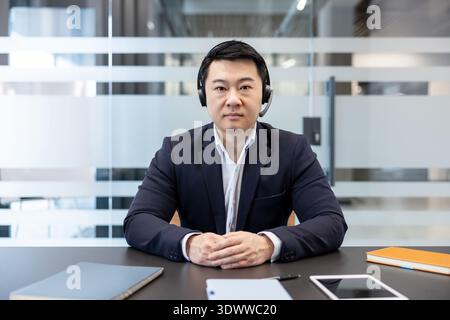 Asiatischer Mann im Anzug, der ein Headset trägt und direkt in die Kamera blickt, an einem virtuellen Meeting teilnimmt oder Kundensupport aus einem modernen Büro bietet Stockfoto