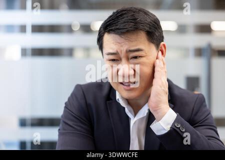 Asiatischer Geschäftsmann, der scharfe Ohrschmerzen oder Unwohlsein erlebt, sein Ohr mit einem gestörten Gesichtsausdruck berührt, was auf ein Gesundheitsproblem in einer Büroumgebung hinweist Stockfoto