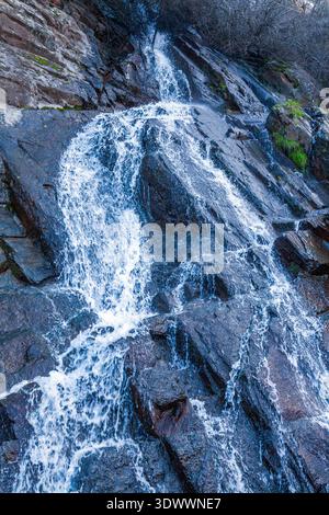 Ein kaskadierender Wasserfall stürzt einen steilen, dunklen felsigen Hang hinunter und erzeugt weißen Schaum, während er über geschichtete Steine mit spärlicher Vegetation fließt Stockfoto