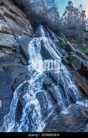 Ein kaskadierender Wasserfall stürzt einen steilen, dunklen felsigen Hang hinunter und erzeugt weißen Schaum, während er über geschichtete Steine mit spärlicher Vegetation fließt Stockfoto