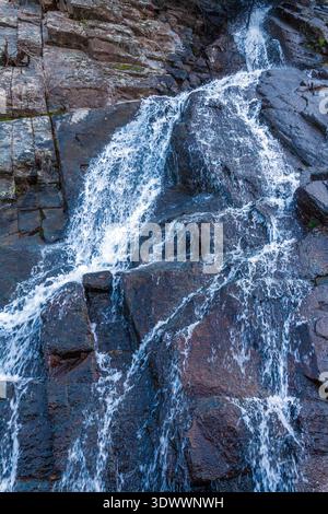 Ein kaskadierender Wasserfall stürzt einen steilen, dunklen felsigen Hang hinunter und erzeugt weißen Schaum, während er über geschichtete Steine mit spärlicher Vegetation fließt Stockfoto