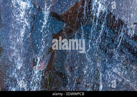 Ein kaskadierender Wasserfall stürzt einen steilen, dunklen felsigen Hang hinunter und erzeugt weißen Schaum, während er über geschichtete Steine mit spärlicher Vegetation fließt Stockfoto