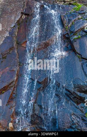 Ein kaskadierender Wasserfall stürzt einen steilen, dunklen felsigen Hang hinunter und erzeugt weißen Schaum, während er über geschichtete Steine mit spärlicher Vegetation fließt Stockfoto