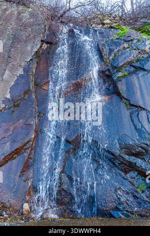 Ein kaskadierender Wasserfall stürzt einen steilen, dunklen felsigen Hang hinunter und erzeugt weißen Schaum, während er über geschichtete Steine mit spärlicher Vegetation fließt Stockfoto