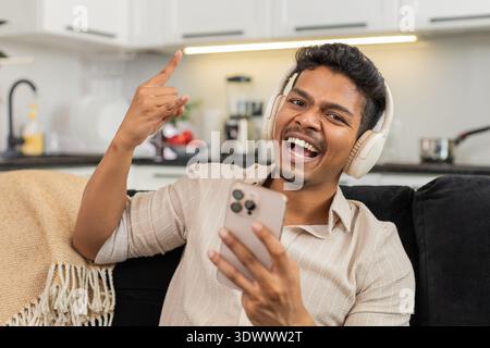 Indischer Mann zu Hause spielt Titel auf dem Smartphone mit schnurlosen Kopfhörern, die zu Beat und Lächeln nicken. Ein Araber auf dem Sofa macht ein Steinschild und schickt Antworten in ch Stockfoto