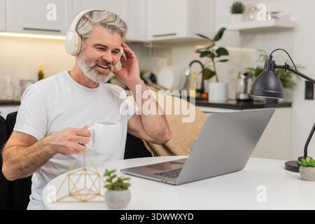 Ein Mann mittleren Alters zu Hause arbeitet an einem Laptop Lächeln hört Musik in Kopfhörern trinkt Kaffee am Tisch. Ein reifer Kerl auf dem Sofa entspannt sich eine Pause von ta Stockfoto