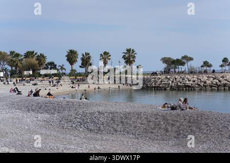 Die Leute entspannen sich am Kiesstrand mit Palmen und felsigem Wellenbrecher unter klarem Himmel. Menton, Frankreich Stockfoto