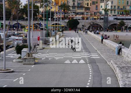 BMX-Fahrer springen in der Luft auf der Uferpromenade mit Palmen und farbenfrohen Gebäuden in der Abenddämmerung. Menton, Frankreich Stockfoto