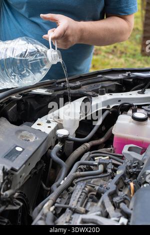 Eine Person gießt Scheibenwaschflüssigkeit aus einer Flasche in den Behälter eines Autos in einer Auffahrt. Die Sonne scheint und hebt die Autos hervor Stockfoto