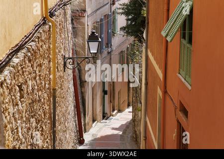 Enge, sonnendurchflutete Gasse mit bunten Fassaden, grünen Fensterläden und schmiedeeisernen Laternen im alten Dorf. Menton, Frankreich Stockfoto