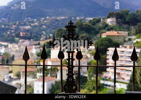 Kunstvoll verziertes schmiedeeisernes Kreuz auf dem Friedhofsgeländer mit Blick auf das Dorf auf dem Hügel. Menton, Frankreich Stockfoto
