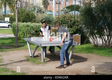 Zwei Männer genießen zwangloses Tischtennis auf einem öffentlichen Tisch im Freien an der palmengesäumten Promenade. Menton, Frankreich Stockfoto