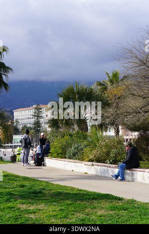 Leute schlendern und entspannen auf der palmengesäumten Strandpromenade unter bewölktem Berghimmel. Menton, Frankreich Stockfoto