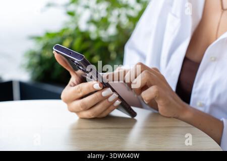 Eine Frau sitzt an einem runden Holztisch in einem Café im Freien und konzentriert sich auf ihr Smartphone. Die Szene spiegelt mobile Kommunikation, digitalen Lifestyle und Re wider Stockfoto